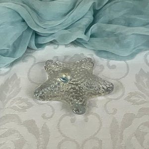 May include: A clear glass starfish ornament with a textured surface. The starfish is sitting on a white tablecloth with a floral pattern.