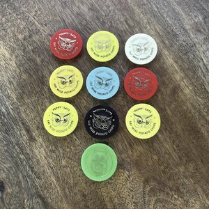 May include: A collection of vintage "Peppy Says Eat Wise Potato Chips" tokens in various colors, including red, yellow, white, blue, and green. Each token features an owl graphic and text. The tokens are arranged on a wooden surface.