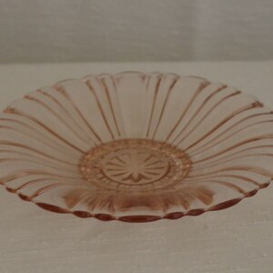 May include: A pink glass bowl with a scalloped edge and a fluted design. The bowl has two small handles on either side.