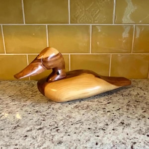 Puede incluir: Un señuelo de pato de madera tallado en madera con un acabado de madera natural. El pato tiene una cabeza marrón y un cuerpo marrón claro.