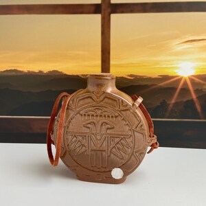 May include: A round, brown ceramic canteen with a cork stopper and a brown leather strap. The front features a raised, embossed design of a stylized bird figure surrounded by geometric patterns. A small white circular sticker is on the lower front.