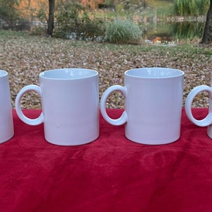 May include: Four white ceramic mugs with handles, lined up on a red surface.