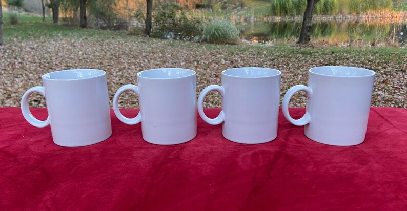 May include: Four white ceramic mugs with handles, lined up on a red surface.