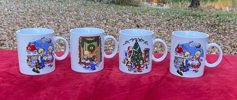 May include: Set of four white ceramic mugs with Christmas scenes. Each mug features a different scene with children and winter imagery. The mugs have a handle on the right side.