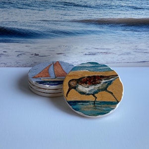 May include: A stack of round coasters with coastal designs. One coaster features a sailboat with an orange sail, and another depicts a shorebird in watercolor. The coasters have a white border and are set against a blurred ocean backdrop.