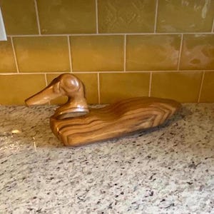 Puede incluir: Un señuelo de pato de madera con un acabado de veta de madera natural. El pato está tallado en un estilo realista con una cabeza y un cuerpo detallados.