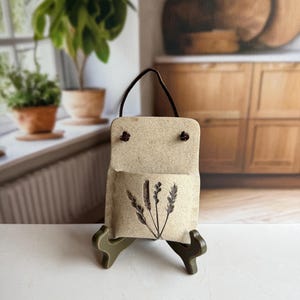 May include: A rectangular, beige ceramic wall pocket with a speckled texture, featuring a dark brown leather hanging cord and a black botanical illustration of three wheat stalks on the front, displayed on a small green plastic stand.
