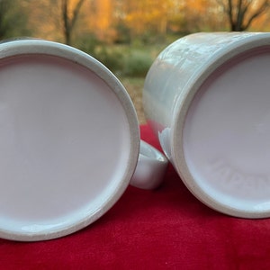 May include: Two white ceramic mugs with a smooth, glossy finish. The mugs are empty and have a light pink interior.