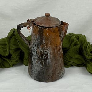 Puede incluir: Una tetera de cerámica marrón rústica con tapa y asa. La tetera tiene un aspecto desgastado y envejecido con una pátina blanca y gris.
