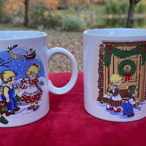 May include: Two white ceramic mugs with Christmas-themed illustrations. One mug features a boy and girl in winter clothes standing in front of a house with a red roof. The other mug shows a boy and girl looking at a decorated door with a wreath.
