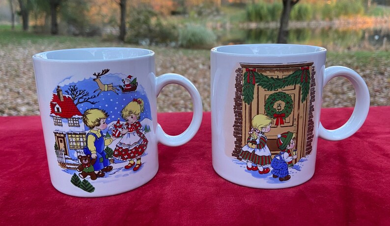 May include: Two white ceramic mugs with Christmas-themed illustrations. One mug features a boy and girl in winter clothes standing in front of a house with a red roof. The other mug shows a boy and girl looking at a decorated door with a wreath.