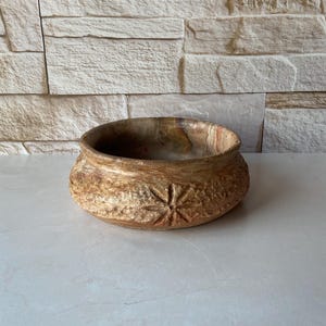May include: A handcrafted, round wooden bowl with a textured exterior and a starburst design. The bowl has a warm, earthy color palette with shades of brown and tan. The interior of the bowl is a darker brown.