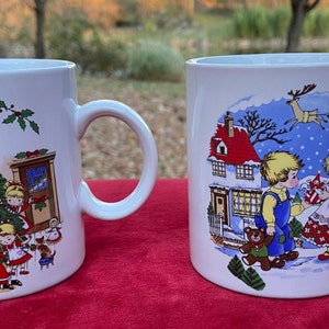 May include: Two white ceramic mugs with Christmas scenes. One mug features a Christmas tree with children decorating it. The other mug shows a couple in a snowy scene with a house in the background.