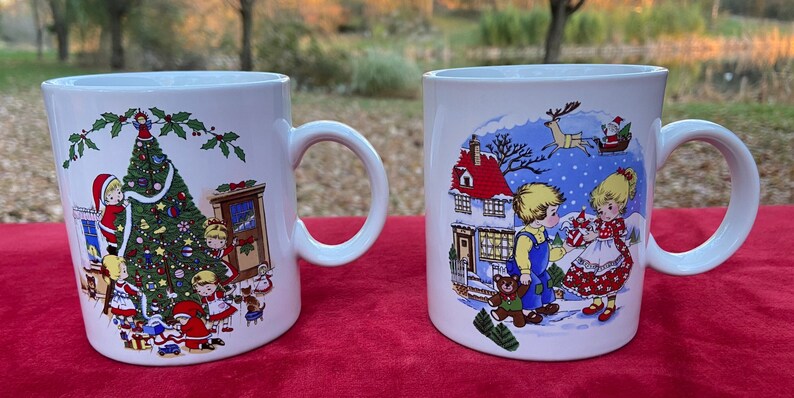 May include: Two white ceramic mugs with Christmas scenes. One mug features a Christmas tree with children decorating it. The other mug shows a couple in a snowy scene with a house in the background.