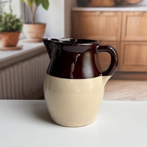 May include: A ceramic pitcher featuring a two-tone design with a glossy dark brown glaze on the upper portion and a smooth cream-colored glaze on the lower body. The vessel includes a curved handle and a small pouring spout at the rim.