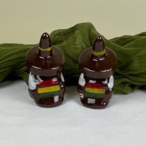 May include: A pair of brown ceramic salt and pepper shakers shaped like people wearing sombreros. The shakers are decorated with colorful stripes and white accents.