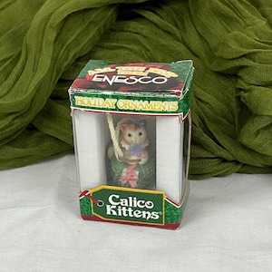 May include: A boxed Calico Kittens holiday ornament featuring a white kitten wearing a pink bow and sitting in a green wreath. The box is red, white, and green and says "All The Best From ENESCO Holiday Ornaments".