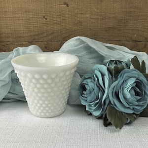 May include: A white hobnail glass vase with a flared top. The vase is surrounded by a light blue fabric and a cluster of teal-colored artificial roses. The background is a wooden surface.
