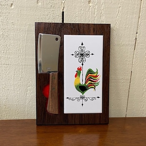 May include: A wooden wall-mounted knife holder with a ceramic tile featuring a rooster design. The tile is white with black accents and a green and yellow rooster with a red comb.