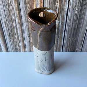 May include: A handmade ceramic vase with a unique design. The top portion is a dark brown color, transitioning to a light gray base with a painted cattail design. The vase has an irregular opening and a small hole near the top.