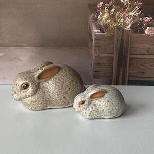 May include: Two ceramic rabbit figurines. The larger rabbit is in the foreground, with a smaller one behind it. Both are speckled gray with brown ears and dark eyes. The background includes a wooden box with flowers.
