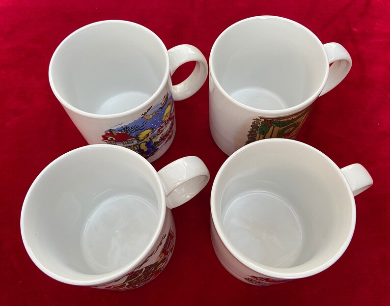 May include: Four white ceramic mugs with colorful holiday-themed illustrations. The mugs have a rounded shape and a handle on the right side.
