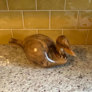 Puede incluir: Una escultura de pato de madera con un acabado de madera natural. El pato está tallado en un estilo realista y tiene un pico y plumas detallados.