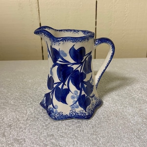 May include: A white ceramic creamer with a blue floral design. The creamer has a scalloped edge and a handle.