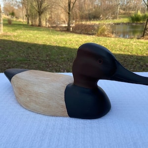 Puede incluir: Un señuelo de pato de madera tallado con cabeza marrón, cuerpo negro y parte inferior marrón claro. El señuelo está pintado para parecerse a un pato real.