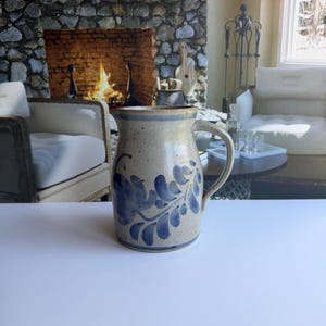 May include: A speckled beige ceramic pitcher featuring a hand-painted blue floral vine pattern. The vessel has a wide, rounded body, a curved handle on the right side, and a flared rim with a small pouring spout at the top center.