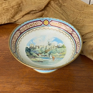 May include: A vintage tin bowl with a blue and white design featuring a picture of Windsor Castle. The bowl has a gold rim and a scalloped edge.