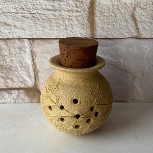 May include: A round, beige ceramic jar with a cork stopper. The jar has a textured surface with leaf-like patterns and small circular holes. The cork is a dark brown color.
