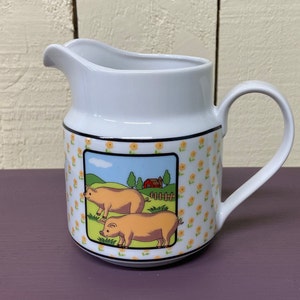 May include: White ceramic pitcher with a handle, featuring a cartoon illustration of two pigs in a farm setting. The pitcher has a yellow and white floral pattern.