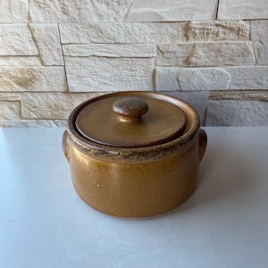 May include: A round, lidded stoneware casserole dish in shades of brown. The lid has a rounded knob and fits snugly on the pot. The dish has two small handles on either side. The stoneware has a speckled, textured appearance.