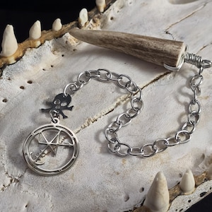 May include: A pendulum featuring a brown, textured antler tip wrapped in silver-toned wire, attached to a silver-toned metal chain. The chain includes a small skull charm and a circular pendant with an intricate, star-like geometric symbol inside.