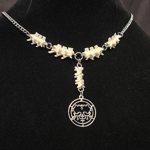 Puede incluir: Un collar con una cadena de metal plateado intercalada con segmentos de vértebras de color blanco hueso. Una sección vertical de tres vértebras se conecta a un colgante circular plateado grabado con un sigilo complejo y simétrico.
