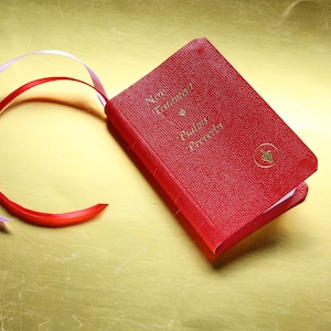 May include: A small, red, textured-cover book titled New Testament, Psalms, and Proverbs in gold lettering. A gold circular emblem is on the front cover. Two thin ribbons, one red and one white, extend from the top of the book spine.