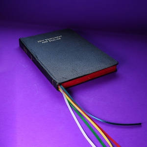 May include: A black, textured, hardcover book titled New Testament and Psalms in gold lettering on the front cover. The book features red-edged pages and four thin ribbon bookmarks in white, green, yellow, and blue extending from the bottom.