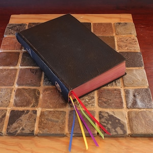 May include: A closed, black leather-bound book with red page edges and colorful ribbon bookmarks. The book is resting on a square, multi-colored tile surface. The ribbons are blue, yellow, purple, green, and red.