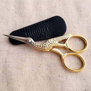 May include: Gold-colored metal embroidery scissors shaped like a crane with a silver-toned pointed tip and a central screw. The scissors are positioned over a black, textured, teardrop-shaped protective sheath with visible stitching.