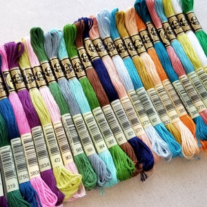 May include: A collection of DMC embroidery floss spools in various colors, including pink, green, blue, brown, and orange. Each spool has a white label with black text indicating the color number and DMC brand name.
