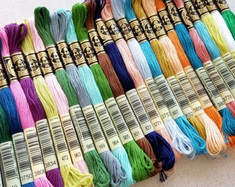 May include: A collection of DMC embroidery floss spools in various colors, including pink, green, blue, brown, and orange. Each spool has a white label with black text indicating the color number and DMC brand name.