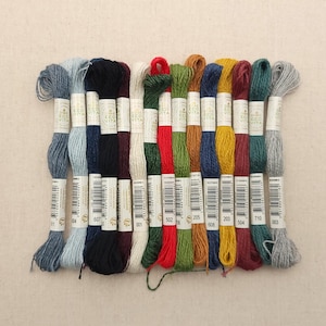 May include: A collection of embroidery floss in various colors, including blue, black, red, green, and yellow. Each skein is labeled with a product code. The floss is arranged neatly, suggesting a craft or sewing project.