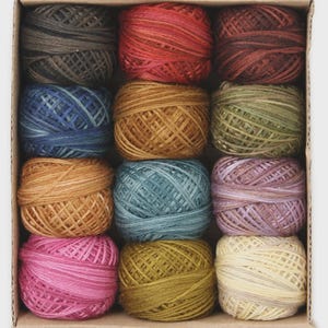 May include: A set of twelve small balls of embroidery thread arranged in a grid inside a square cardboard box. The thread colors include shades of charcoal, red, brown, blue, gold, olive green, tan, teal, purple, pink, mustard, and cream.