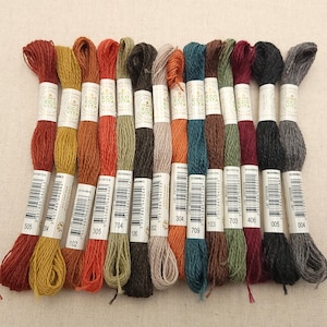 May include: A collection of colorful embroidery floss skeins, each with a paper label. The floss colors range from warm reds and oranges to cool greens and blues. The labels display the brand name and product codes.