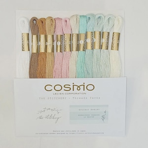 May include: A collection of twelve embroidery thread skeins in a palette of white, tan, brown, pink, and light blue, each with a gold Cosmo label. The threads are mounted on a white card labeled Cosmo Lecien Corporation The Stitchery Threads Packs.