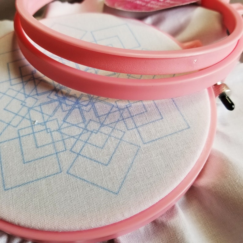 May include: Pink embroidery hoop with a white fabric stretched across it. The fabric has a blue geometric pattern of squares.