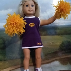 May include: A doll dressed as a cheerleader, wearing a purple and white uniform with the letters "LSU". The doll has blonde hair and is holding two orange pom-poms. The doll is wearing white sneakers with purple accents.