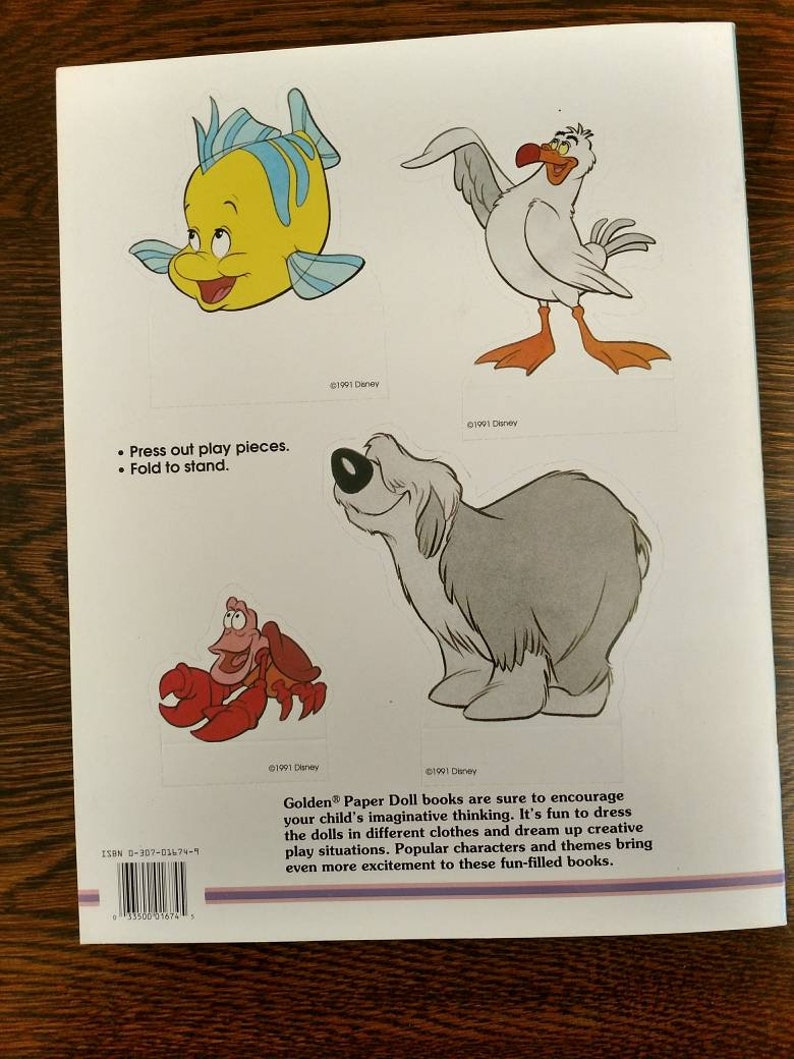 May include: A book cover featuring a colourful illustration of a seagull, a fish, a crab, and a dog. The book is titled "Golden Paper Doll" and includes the text "It's fun to dress the dolls in different clothes and dream up creative play situations."