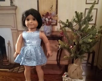 May include: A doll wearing a light blue sequined dress and silver shoes stands next to a small decorated Christmas tree. The doll has dark hair and is posed in a room with a fireplace and antique furniture. The tree is in a burlap sack.
